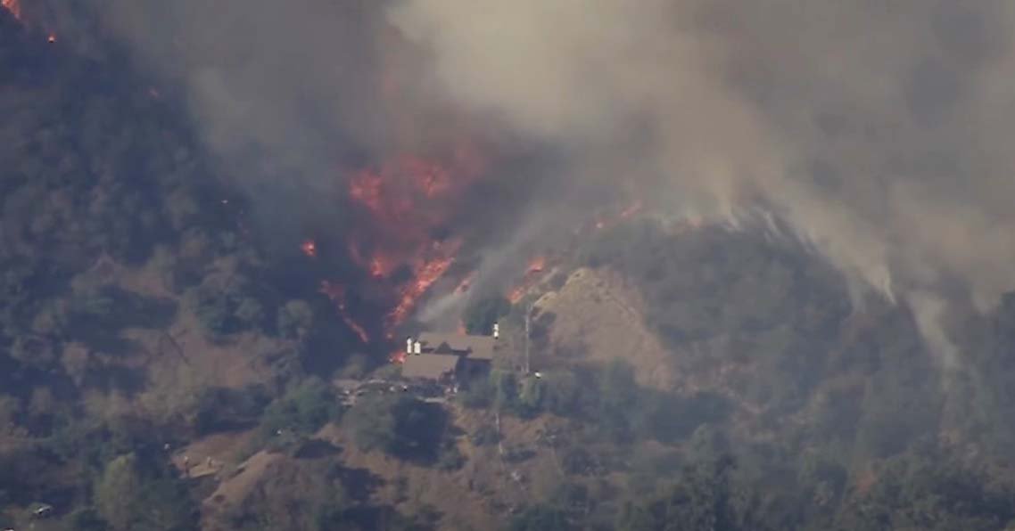 A massive wildfire burning in Los Angeles, viewed from a mountain, with large flames and thick smoke billowing into the sky.
